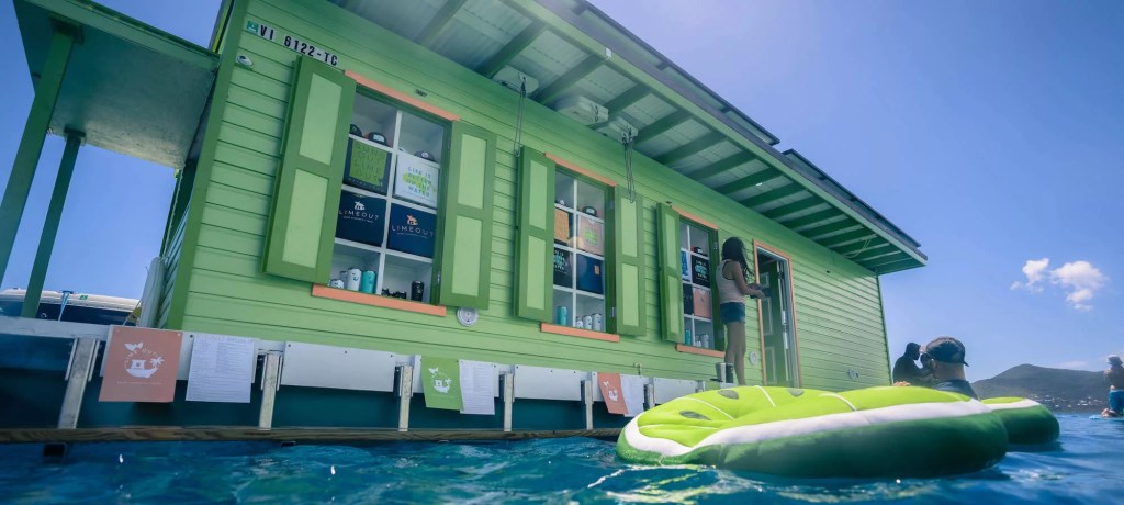 Green floating house on water with person standing at window, inflatable float in foreground.