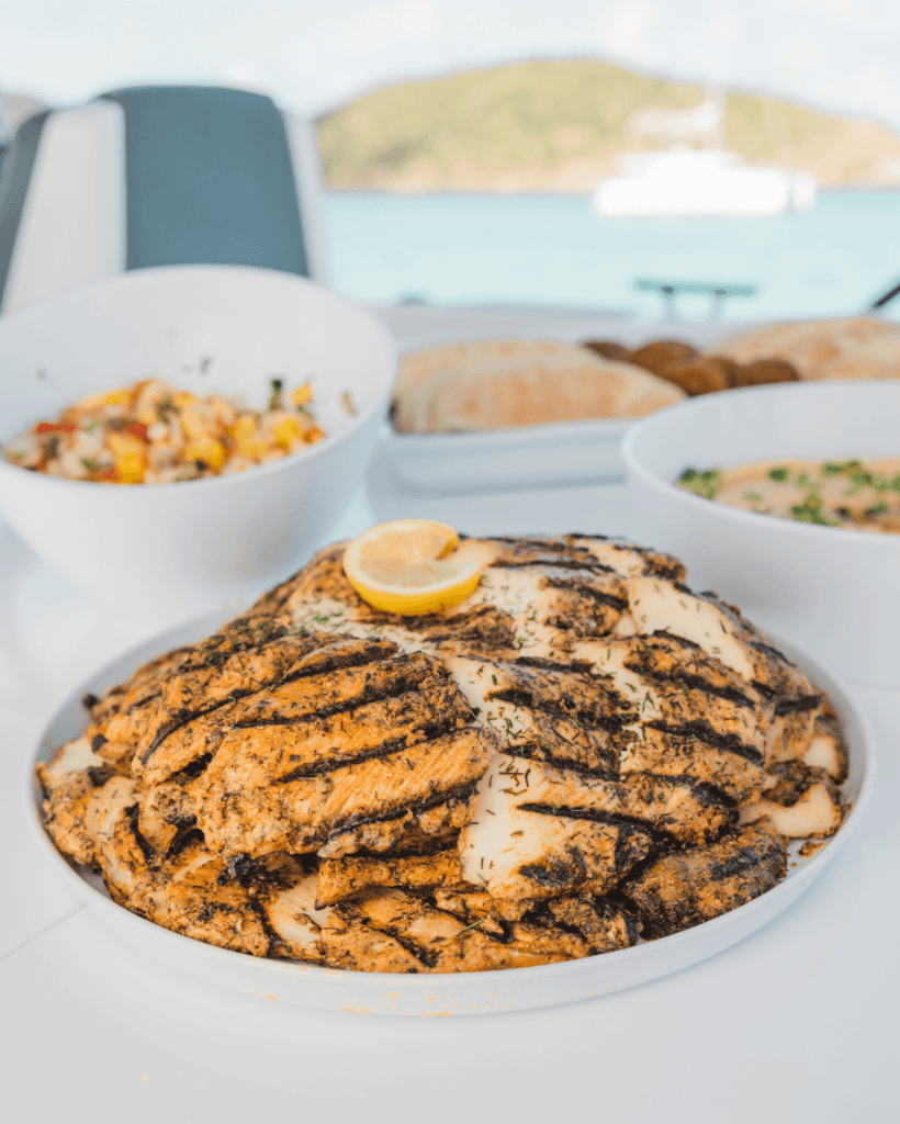 Grilled chicken with lemon garnish on a plate, bowls of food in the background.