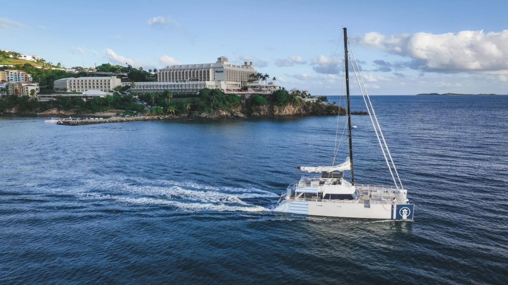 Catamaran sailing near a coastal resort with blue sky and calm sea.